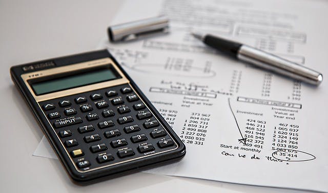 Stock image of a calculator lying next to a sheet of figures with some circled in pen. An uncapped pen lies on top of the papers.